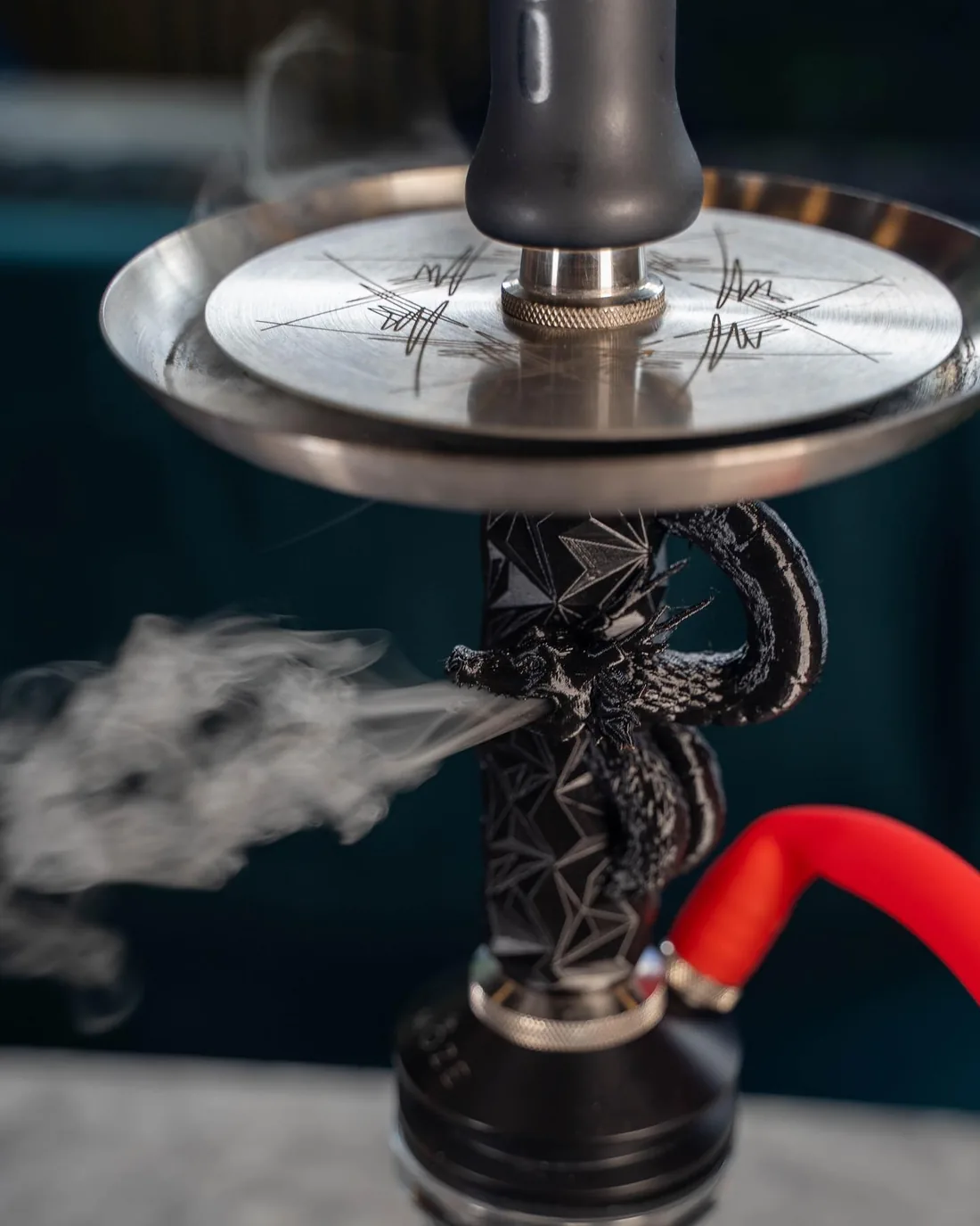 A shisha stem wrapped by a cast black dragon, smoke pouring from its mouth beneath a brushed steel tray, with a red hose resting alongside