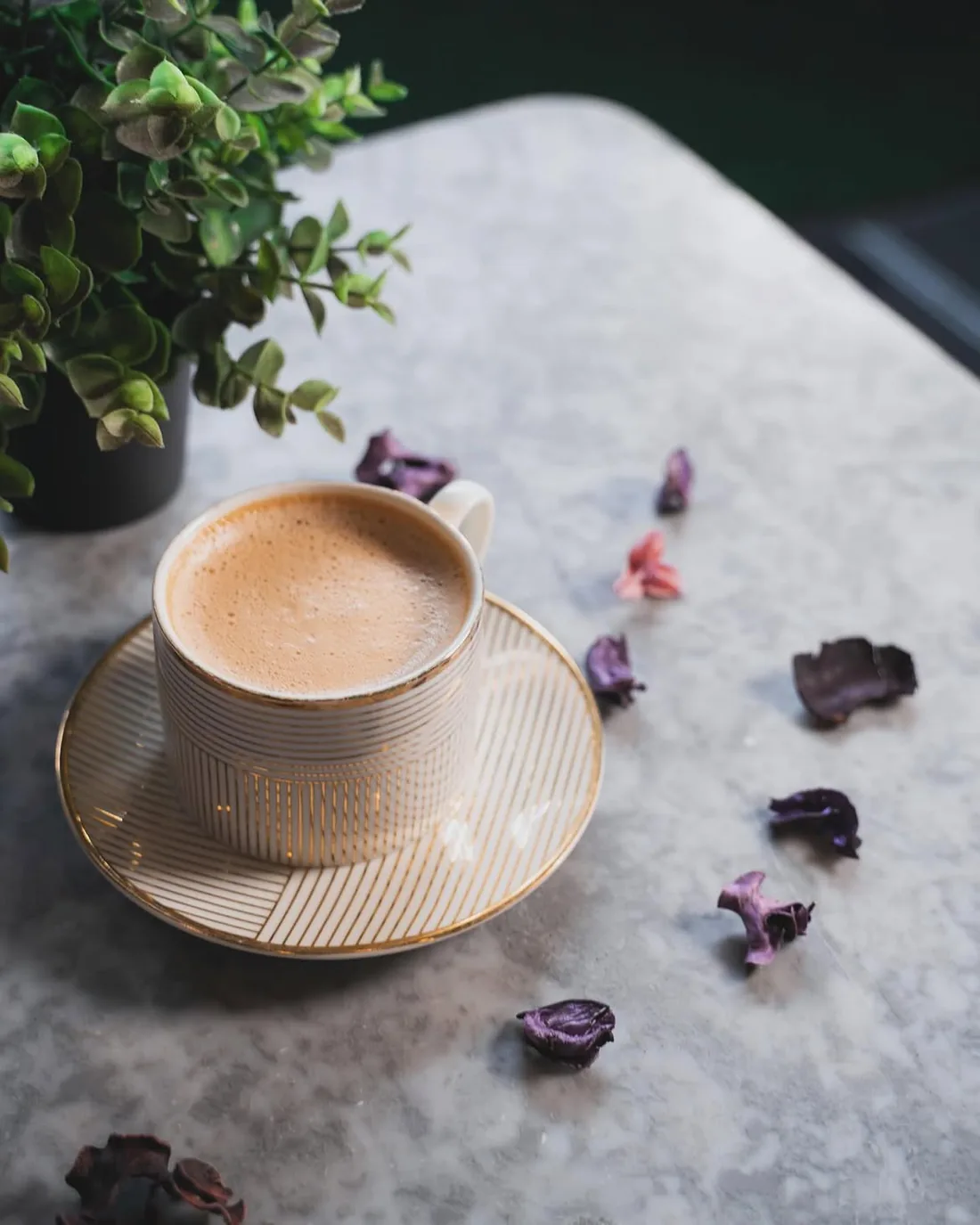 Karak chai in a ribbed cream and gold cup on a matching saucer, set on a marble table beside a small plant and scattered dried petals