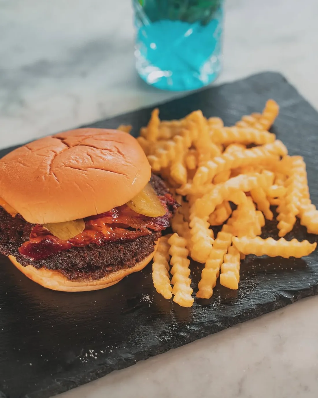 The Smokey Smash burger with beef rashers, melted cheese and sauce in a toasted potato bun, served on a slate board with crinkle-cut fries and a blue mocktail behind
